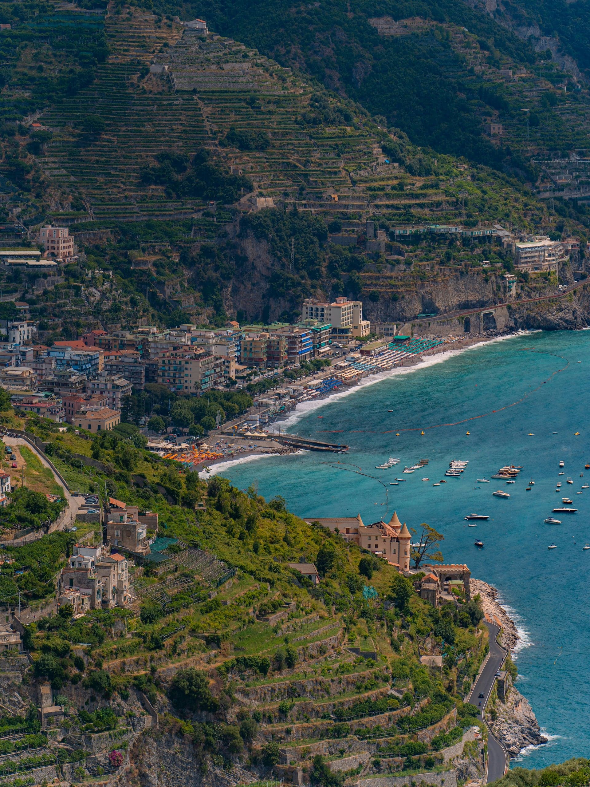 Terraced green hills, a crescent beach, and small boats scattered across blue water below Ravello
