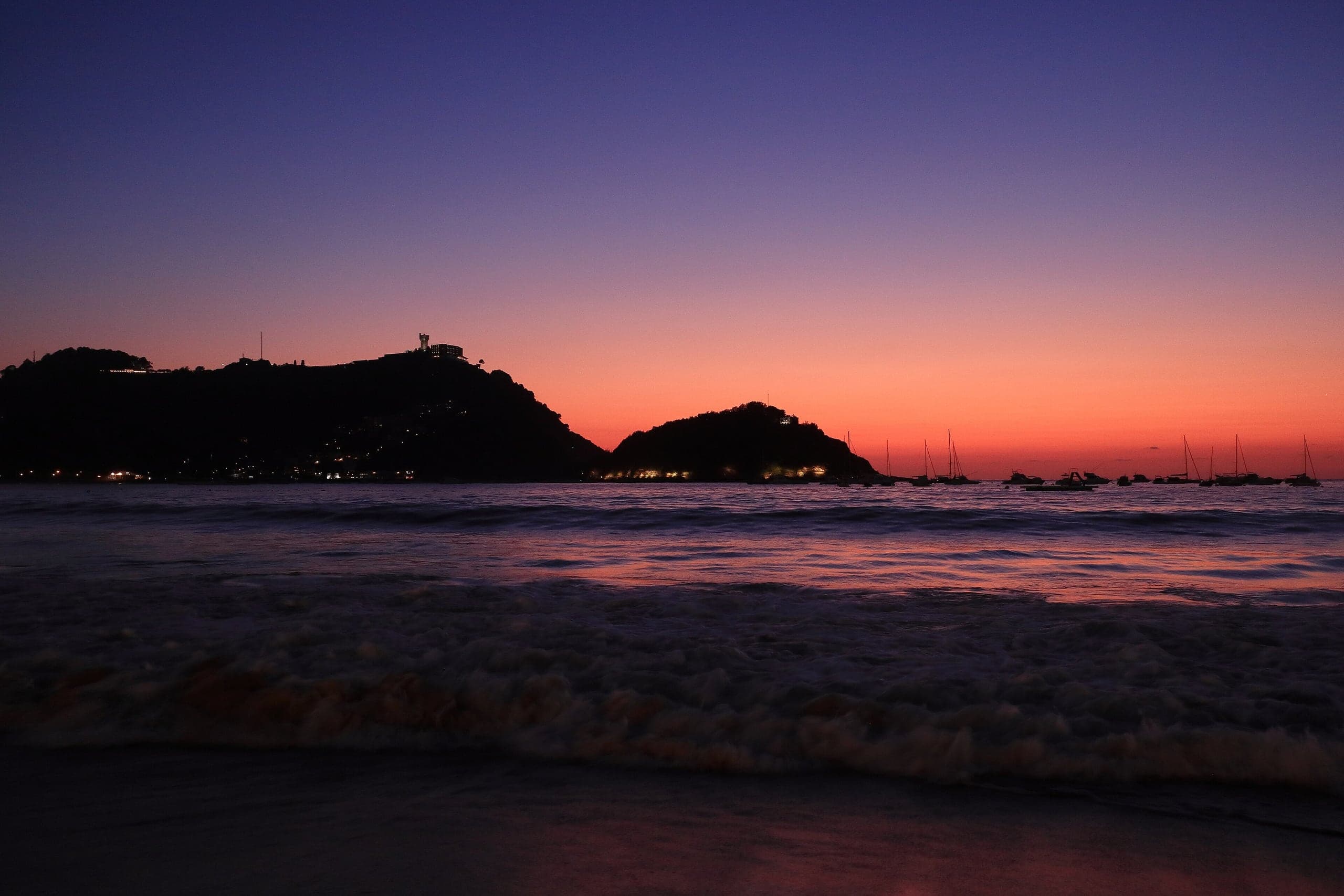 Sailboats and the islands of La Concha Bay silhouetted beneath a deep coral Basque sunset