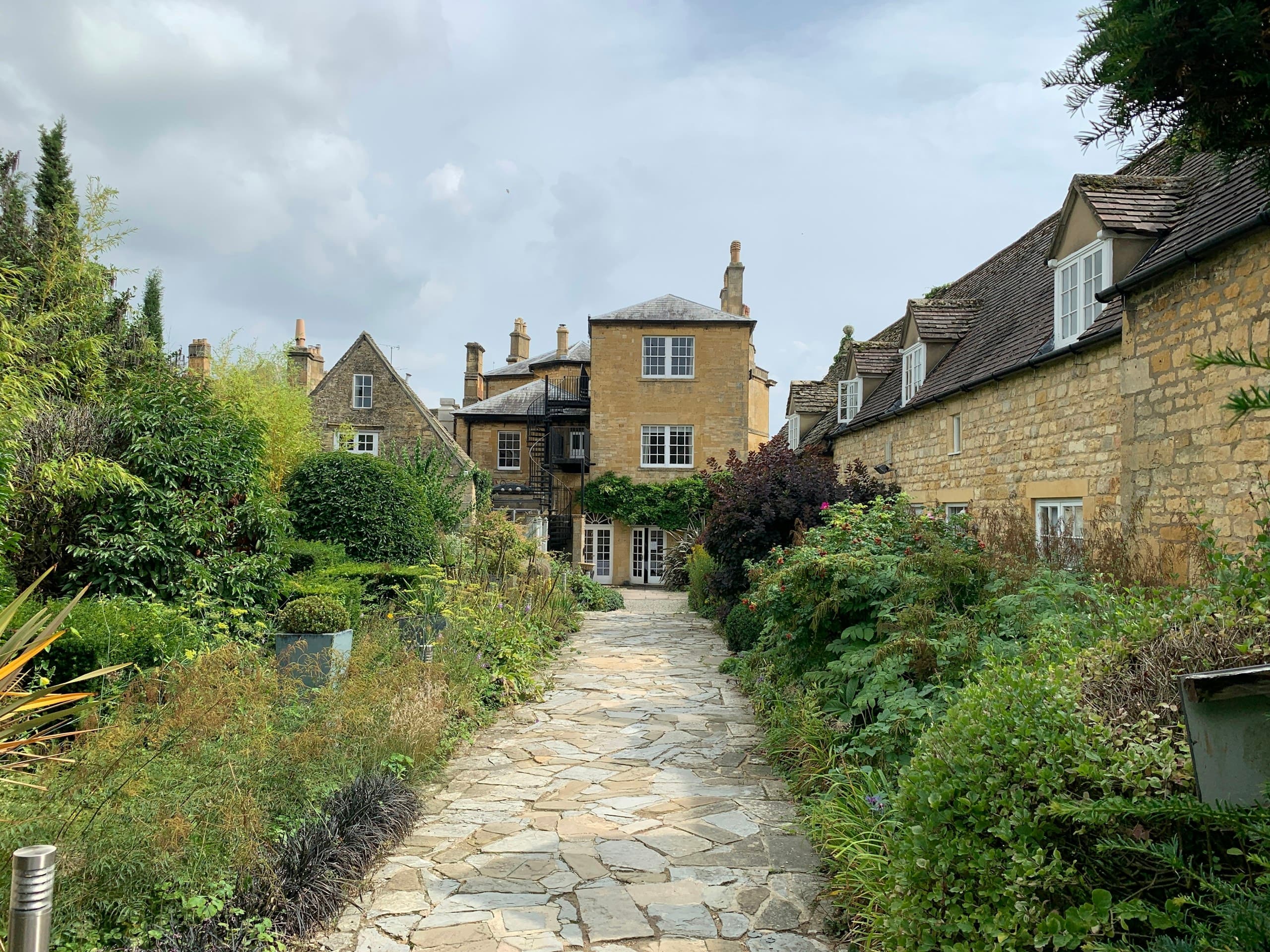 brown brick pathway between green plants and brown concrete houses under white clouds and blue sky