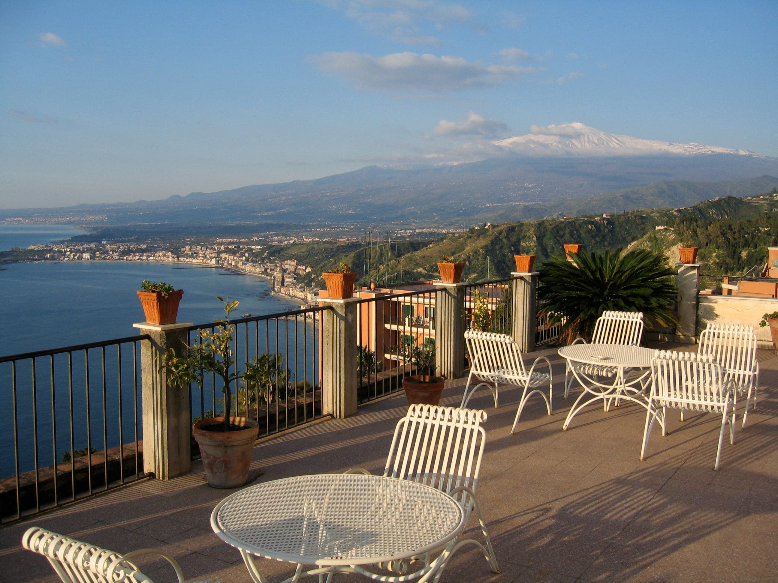 A flower-edged terrace overlooking Taormina's coast and distant Mount Etna beneath a clear Sicilian sky