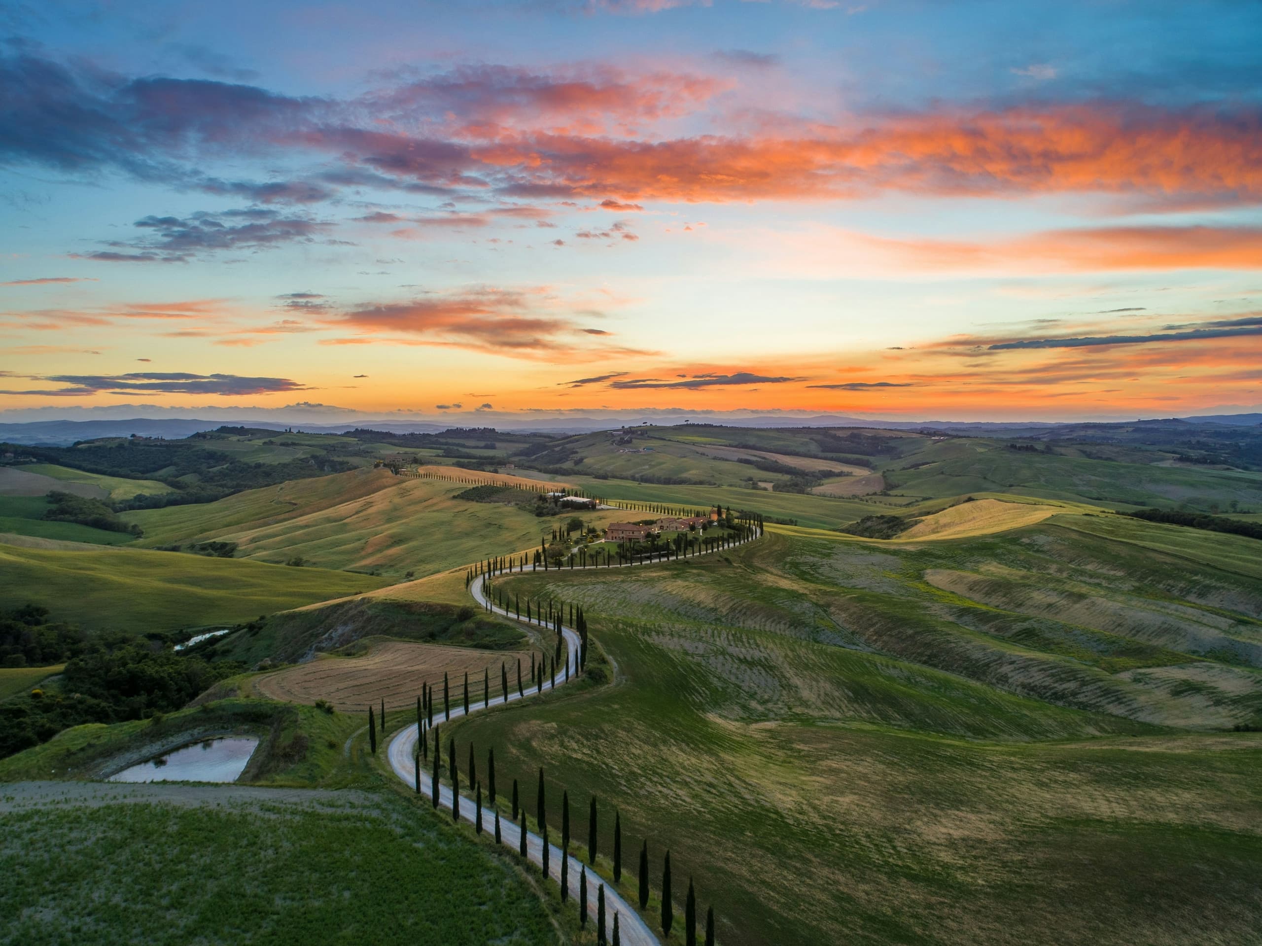 A winding country road crossing green Tuscan hills beneath a luminous gold-and-blue sky