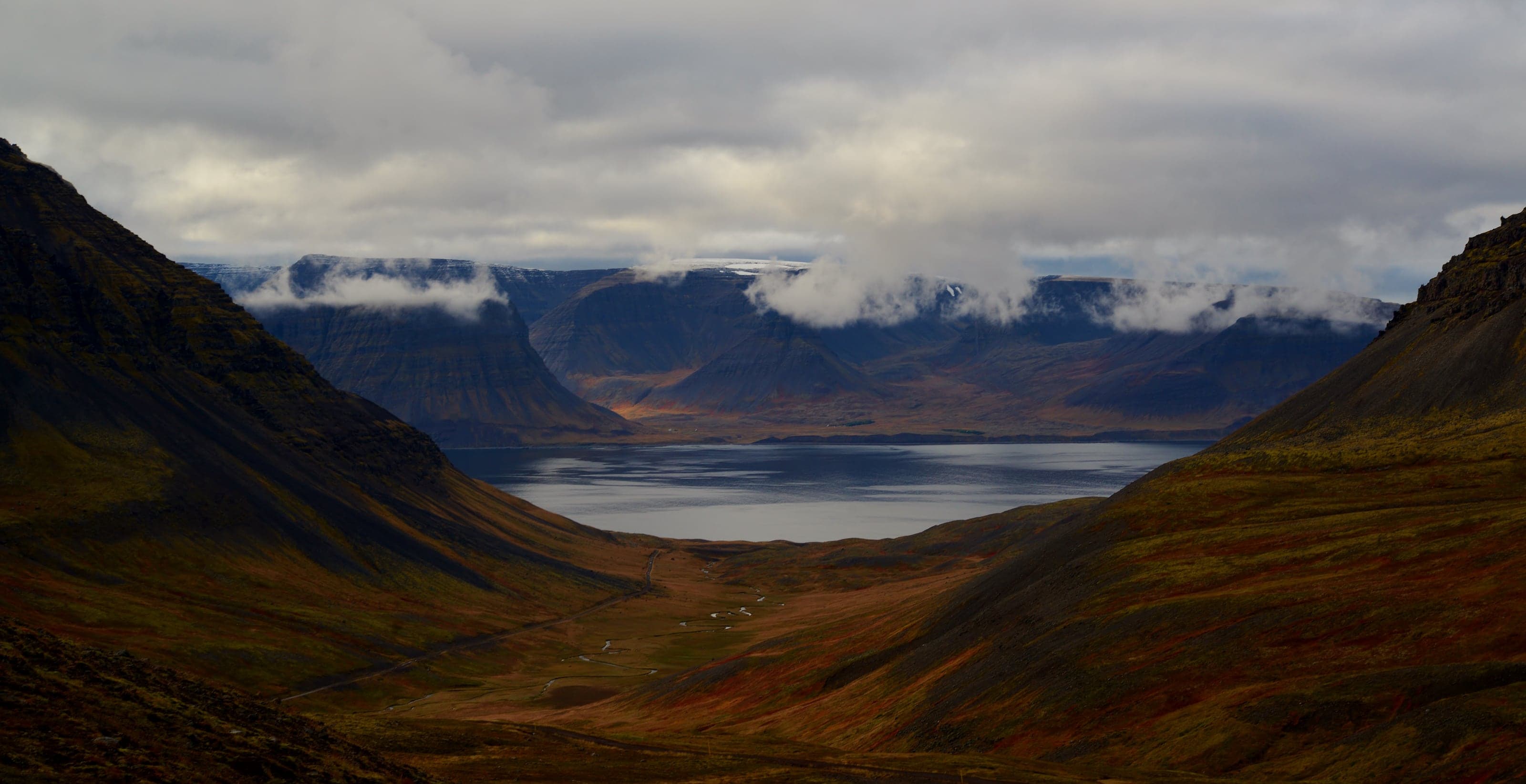 Clouds spilling over dark table mountains into a rust-colored valley and still fjord in western Iceland